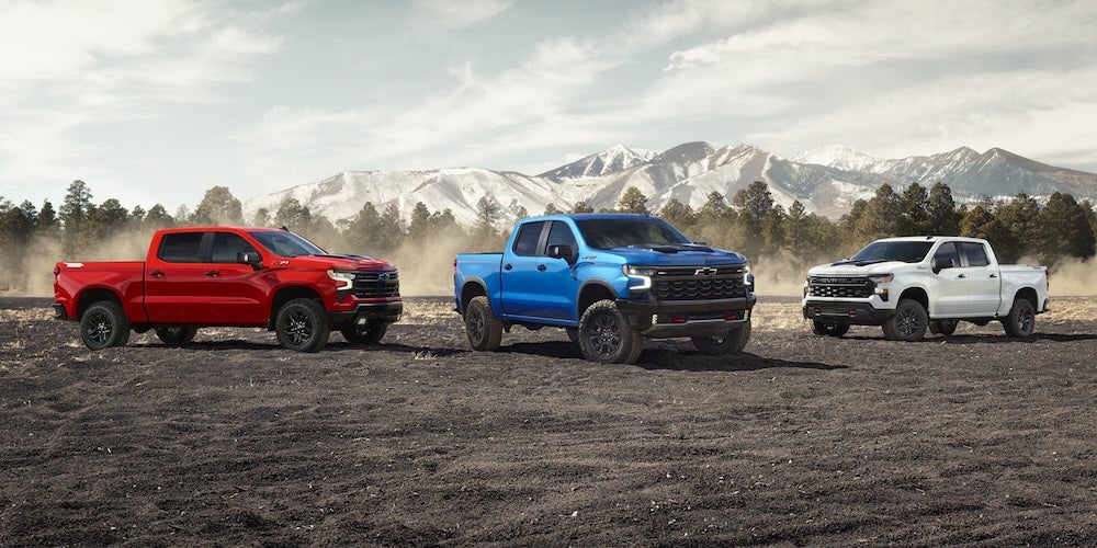 Three Chevrolet Silverado trucks, one red, one blue, and one white, parked on a dirt field with mountains in the background